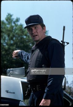 a man in uniform pointing at something while standing next to a car with his hand on the hood