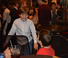 two young boys sitting at a table with drinks in front of them and people standing around