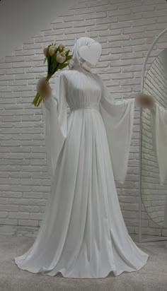 a woman in white dress holding flowers next to a mirror with brick wall behind her