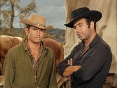 two men standing next to each other in front of some horses and one is wearing a cowboy hat