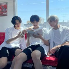 three young men sitting on a red bench looking at a book while another man sits next to them