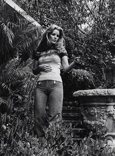 black and white photograph of woman leaning on stone wall in front of trees, bushes and shrubbery