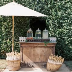 an umbrella and some drinks on a table in front of a bush with bushes behind it