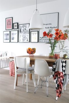 a dining room table with white chairs and pictures on the wall above it, along with vases filled with orange flowers