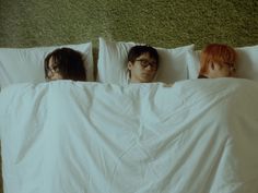 three people laying in bed with white sheets and pillows on top of them, looking at the camera