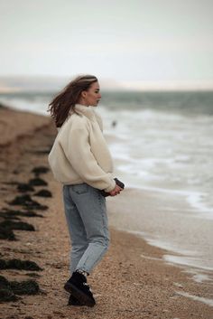 a woman standing on top of a sandy beach next to the ocean with her hands in her pockets