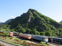 there are many trains on the tracks in front of a large mountain with green trees