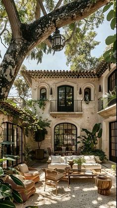 an outdoor living area with couches and tables under a tree in front of a house