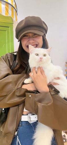 a woman is holding a white cat in her arms and smiling at the camera while wearing a brown leather jacket