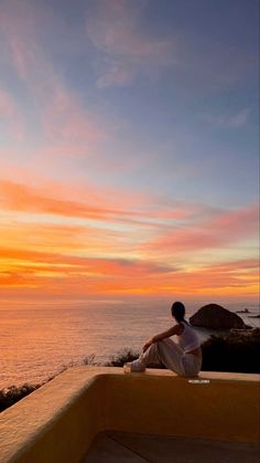 a person sitting on a ledge looking out at the ocean and sunset in the distance