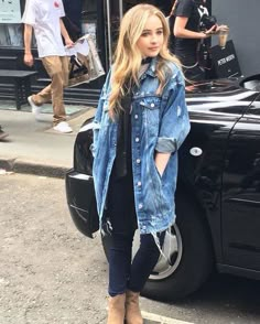 a woman standing in front of a black car wearing a jean jacket and jeans on the street