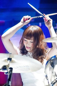 a woman in white dress playing drums with her hands behind her head and holding two sticks above her head