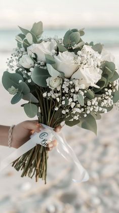 a person holding a bouquet of white flowers and greenery in their hand on the beach