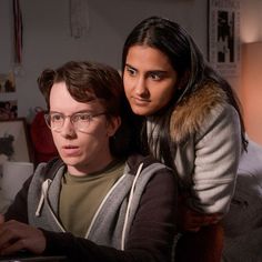 two people sitting in front of a laptop computer on top of a bed, one person wearing glasses and the other looking at the camera