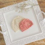 two knitted hats sitting on top of a white doily next to a plant