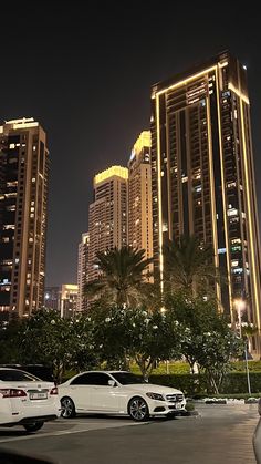 two white cars are parked in front of some tall buildings with lights on at night