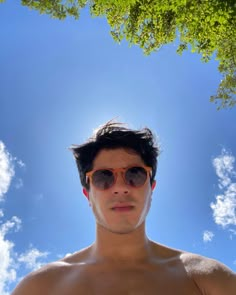 a shirtless man wearing sunglasses under a tree with blue sky and clouds in the background