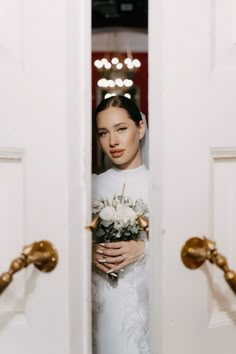 a woman in a white dress is holding a bouquet and looking through the door to another room