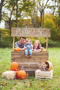 two adults and a child are sitting in a crate with pumpkins on it, surrounded by fall foliage