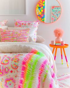 a bed covered in lots of colorful pillows and blankets next to a table with a mirror on it