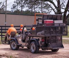 two men in orange shirts are loading items onto a utility vehicle that is parked near a fence