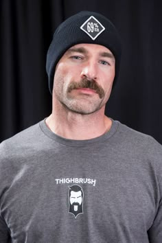 a man with a beard wearing a black hat and grey t - shirt is looking at the camera