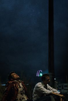 two men sitting on the ground at night with their eyes closed and one looking up