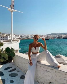 a woman sitting on a wall next to the ocean wearing sunglasses and a white dress