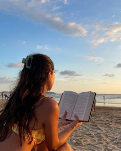 a woman sitting on the beach holding an open book in her hand and reading it