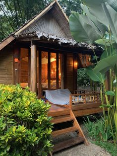 a small wooden cabin in the middle of some trees and bushes with a hammock hanging from it's roof