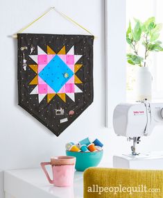 a quilt hanging on the wall next to a sewing machine and bowl with fruit in it