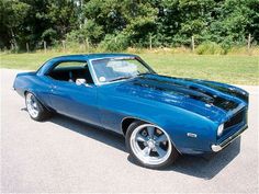 a blue muscle car parked in a parking lot next to some grass and trees on the other side of the road