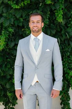 a man in a gray suit and white shirt standing next to a green bush with his hands in his pockets