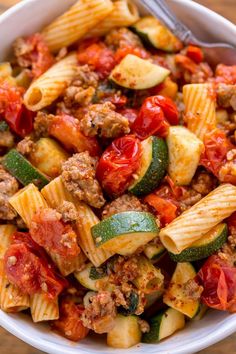 a white bowl filled with pasta and meat on top of a wooden table next to a fork