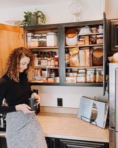 a woman standing in a kitchen holding a coffee mug and looking at the contents on the shelves