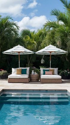 two lounge chairs with umbrellas next to a swimming pool surrounded by palm trees and flowers