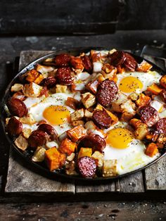 a skillet with eggs, sausage and potatoes on it sitting on a wooden table