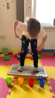 a little boy standing on top of a play mat with cups and toys in front of him