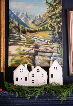 three white houses sitting on top of a mantle next to a painting and two framed pictures