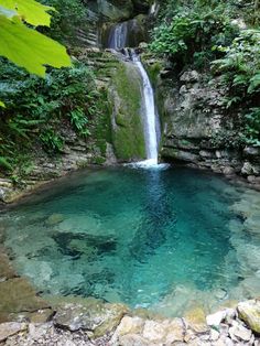 there is a small waterfall in the middle of this pool with clear blue water and green vegetation surrounding it