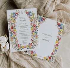 two wedding cards with flowers on them sitting next to a flowery cloth draped fabric