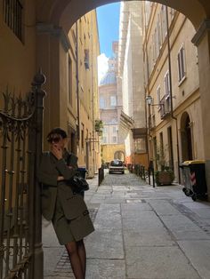 a woman leaning against a gate in an alleyway with buildings on either side and cars parked along the street