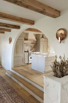 a kitchen with white walls and wooden floors, an archway leading to the living room