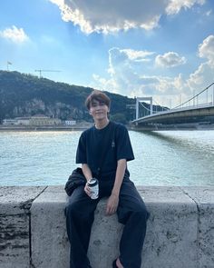 a young man sitting on the edge of a concrete wall next to water with a bridge in the background