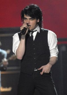 a man in a vest and tie holding a microphone up to his mouth while standing on stage