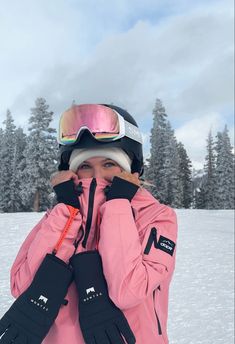 a woman in pink jacket and ski goggles covering her face with both hands while standing on snow covered ground