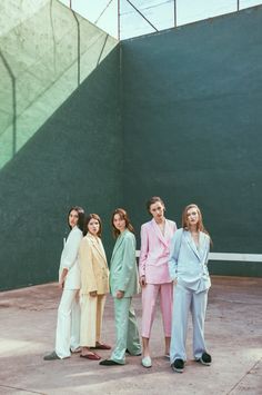 four women standing next to each other in front of a green and white wall, wearing paste colored suits