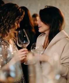 two women standing next to each other with wine glasses in their hands and smiling at each other