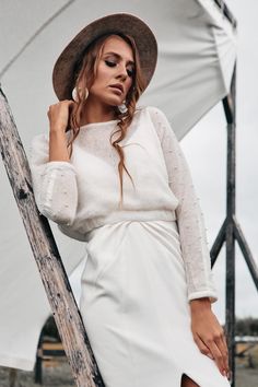 a woman in a white dress and hat leaning against a wooden pole with her hand on her hip