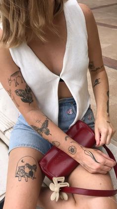 a woman sitting on the ground with tattoos on her arms and legs, holding a red purse